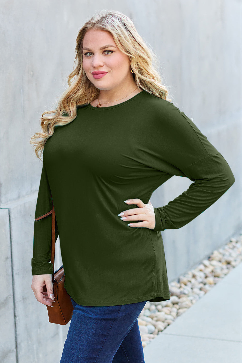 A woman with shoulder-length hair wearing a Basic Bae Full Size Round Neck Dropped Shoulder T-Shirt in green and a pair of jeans, exuding a basic style, stands against a concrete wall. She's holding an imported brown woven backpack on her shoulder while touching her hair with her hand.