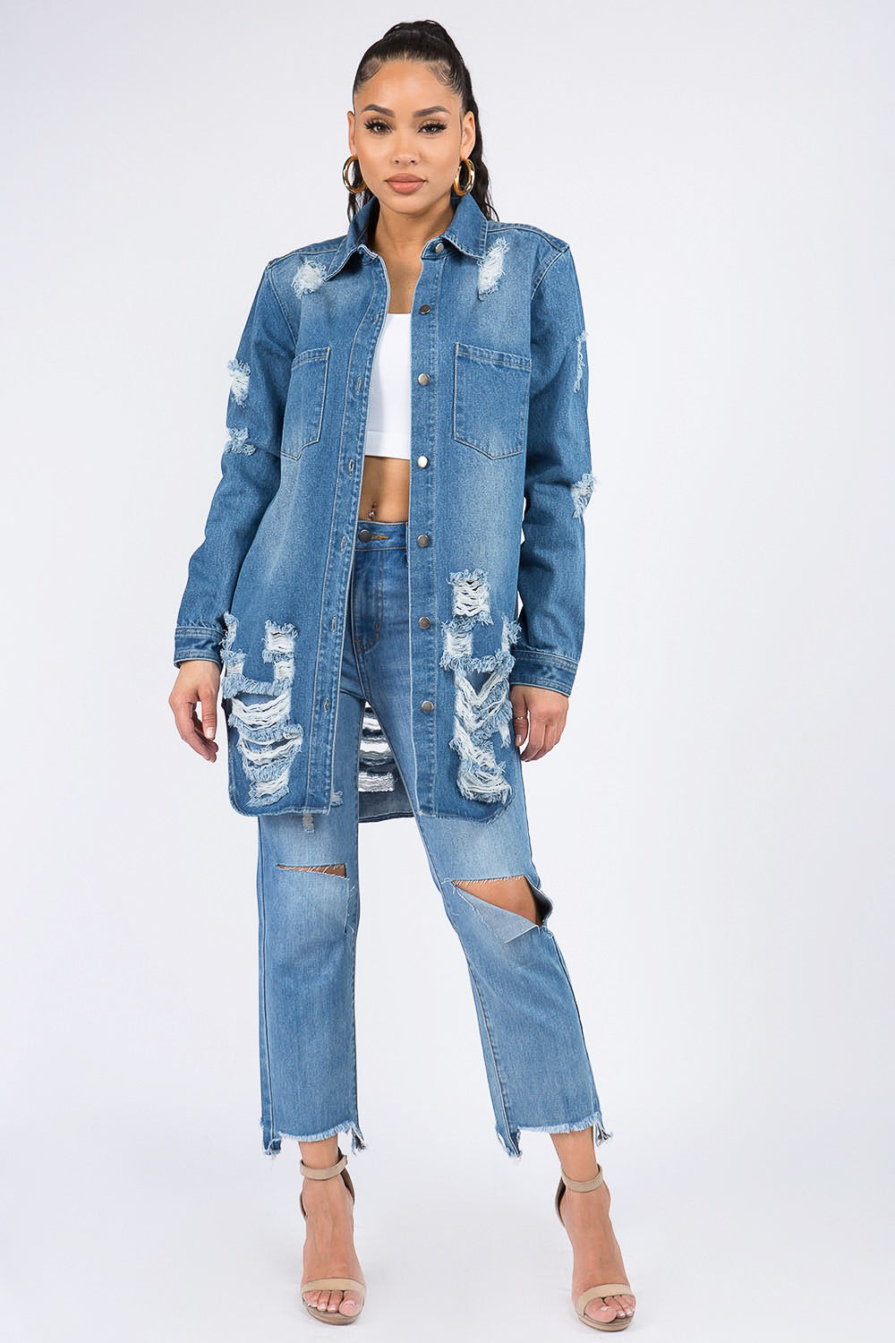 A woman wearing the American Bazi Distressed Button Down Denim Shirt Jacket and jeans paired with a white crop top exudes vintage-inspired charm as she poses with one hand on her head against a plain background.