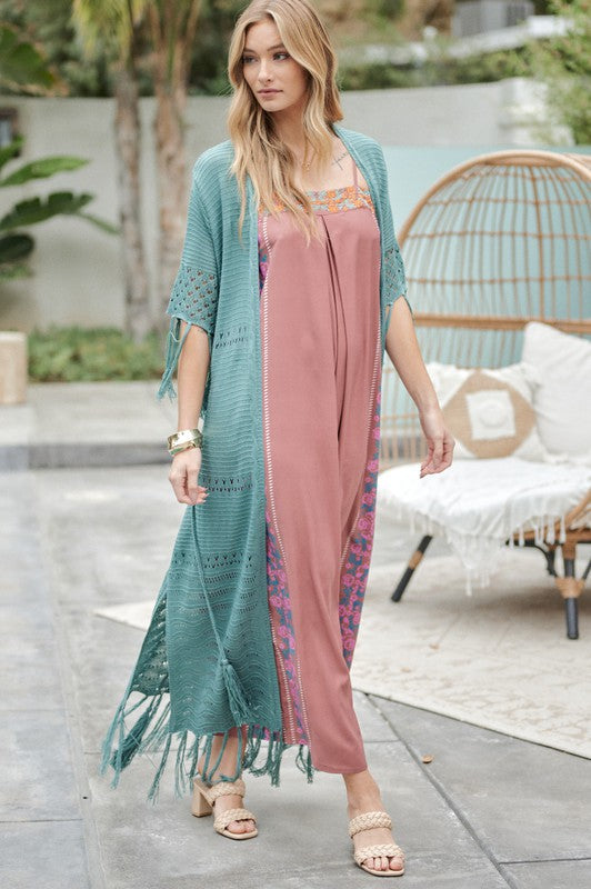 A woman in a green floral dress and a Solid Long Cardigan With Fringe exudes boho-chic vibes as she strolls on a patio, surrounded by outdoor furniture and lush plants.