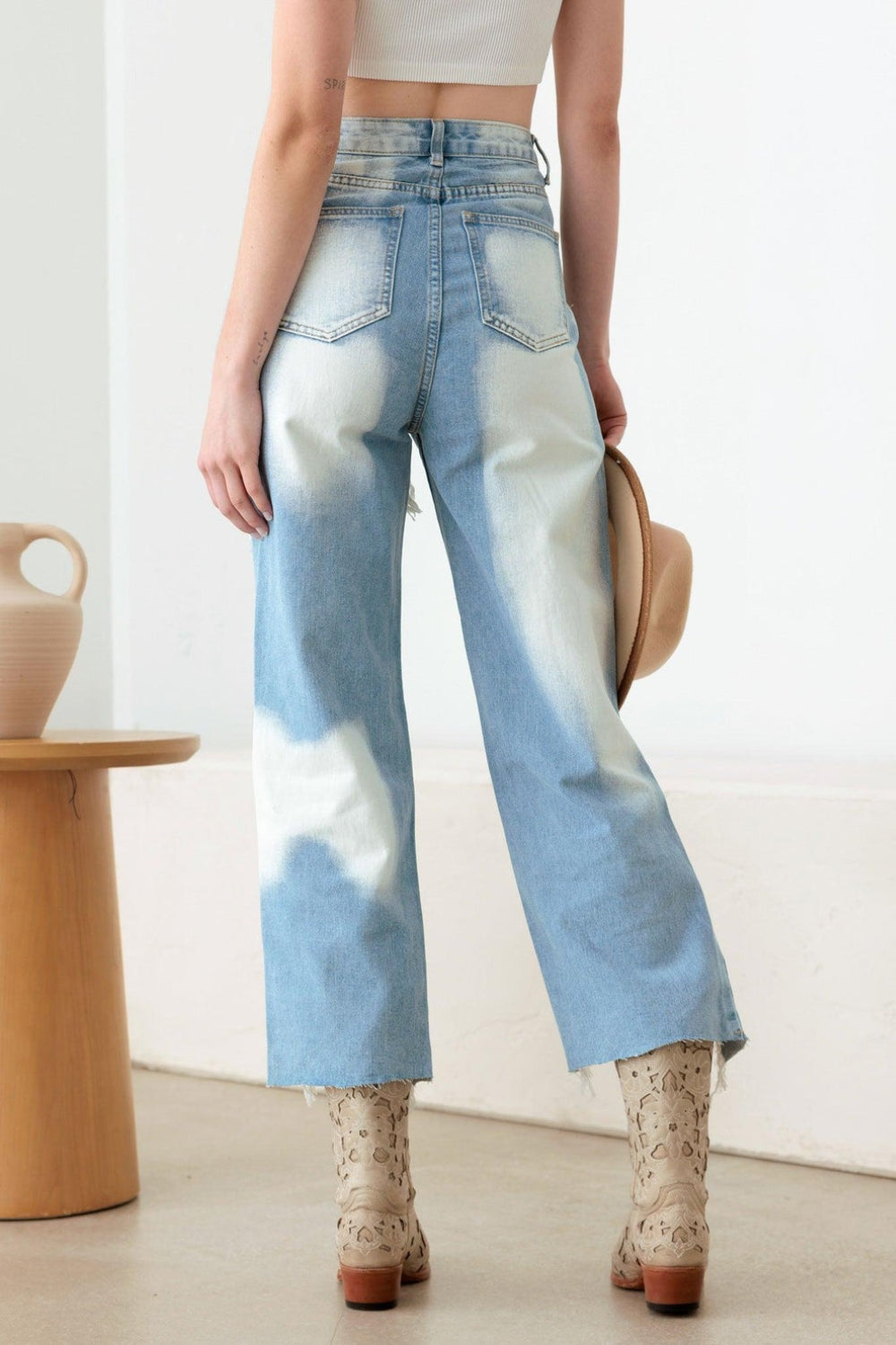 A woman stands indoors in a Western Boho style, showcasing a white crop top and plaid shirt paired with the Litz La Frayed Cut Distressed Jeans. She holds a hat in her hand and completes her outfit with patterned boots, while a vase is positioned on a wooden stool nearby.