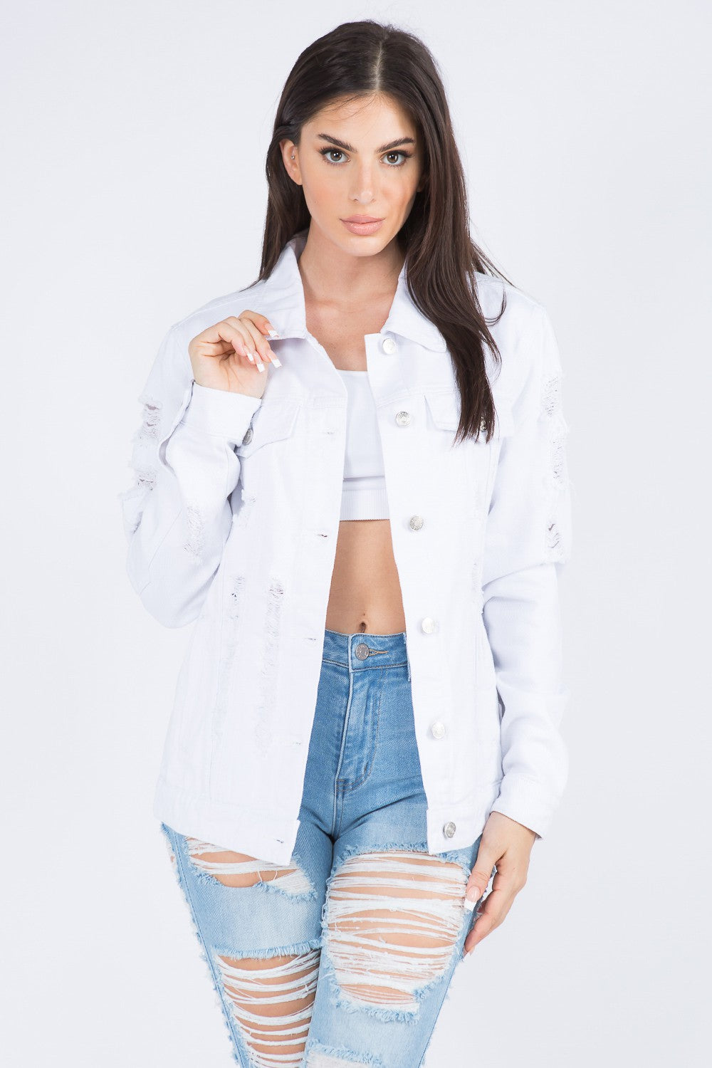 A woman wearing an American Bazi Distressed Back Button Down Denim Jacket over a white crop top and distressed blue jeans stands with her left hand holding the jacket collar.