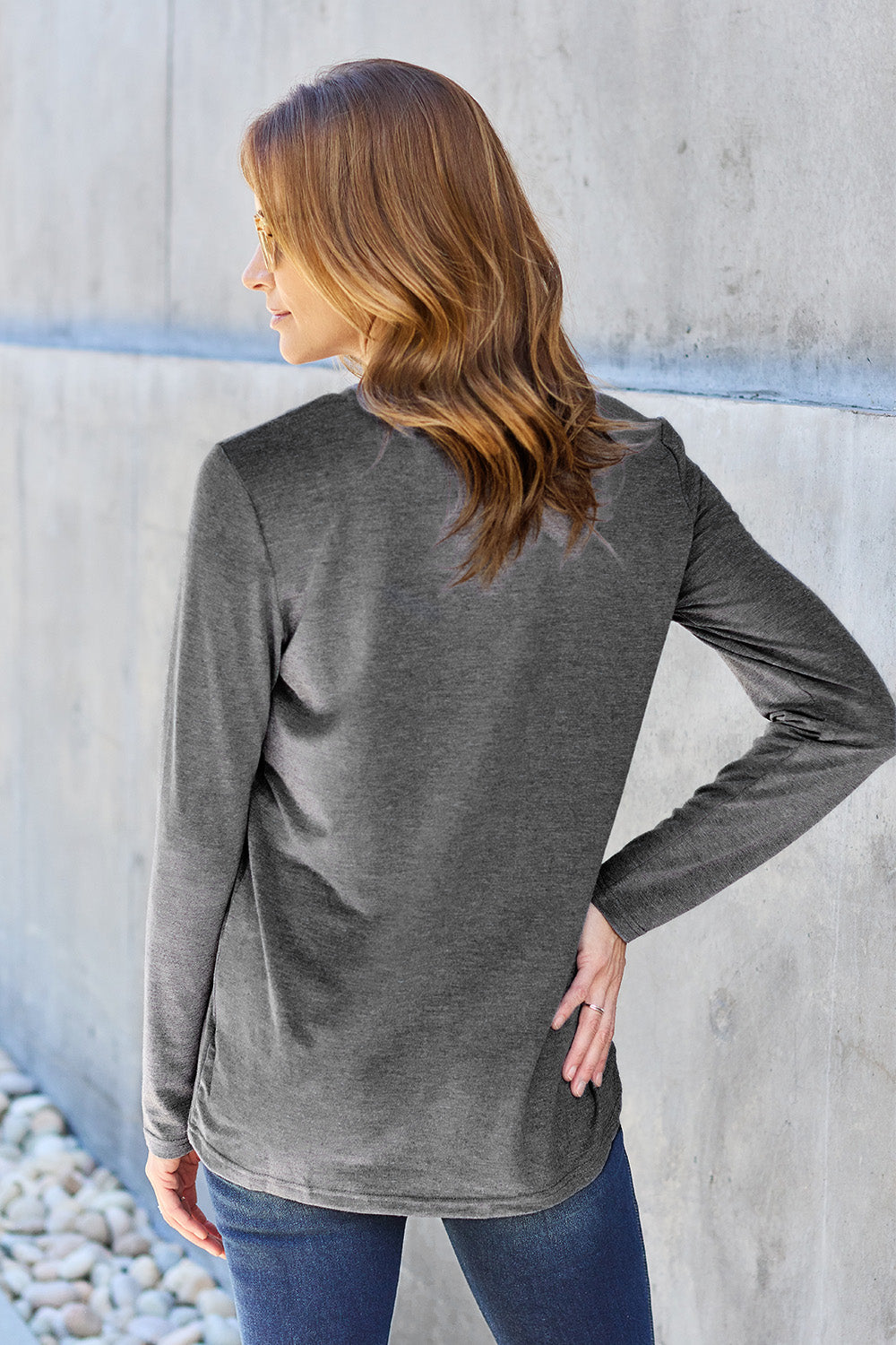 A woman with shoulder-length hair wearing the Basic Bae Full Size Round Neck Long Sleeve Top and blue jeans stands in front of a concrete wall, holding a beige handbag over her shoulder. Her outfit exemplifies basic style, with the top made from 100% polyester and recommended for machine wash cold.