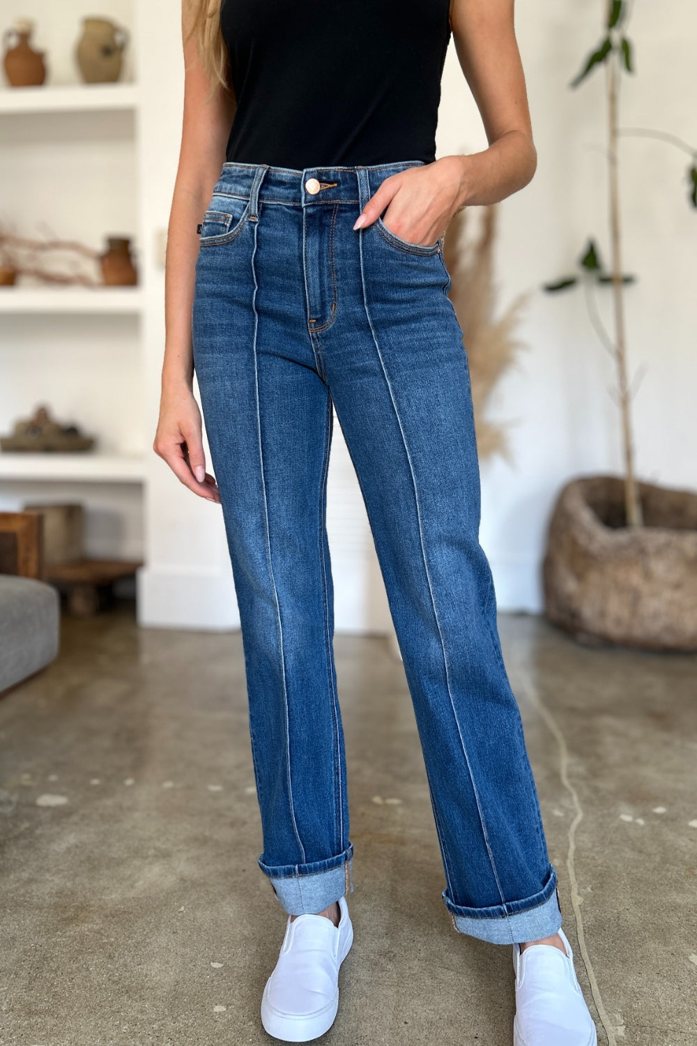 Two women stand side by side indoors, both wearing black tank tops, Judy Blue Full Size High Waist Front Seam Detail Straight Jeans, and white shoes. They are smiling and posing for the camera. A plant and shelves with decorations are in the background.