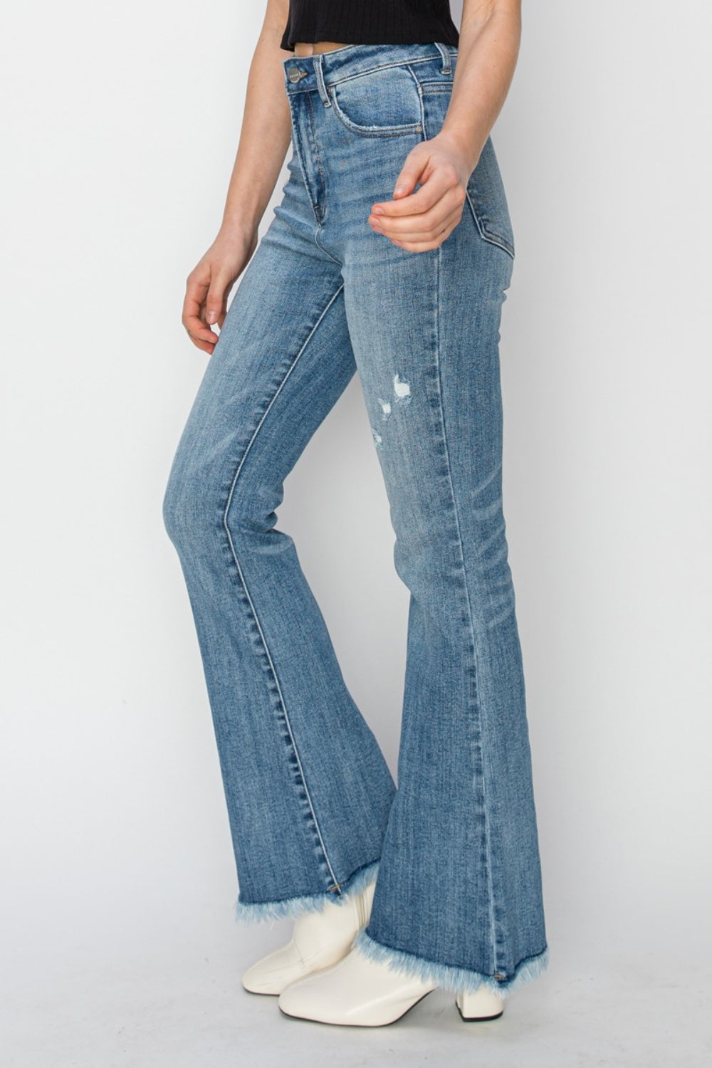 Woman wearing RISEN High Rise Frayed Hem Bootcut Jeans and a black top, standing against a plain background.