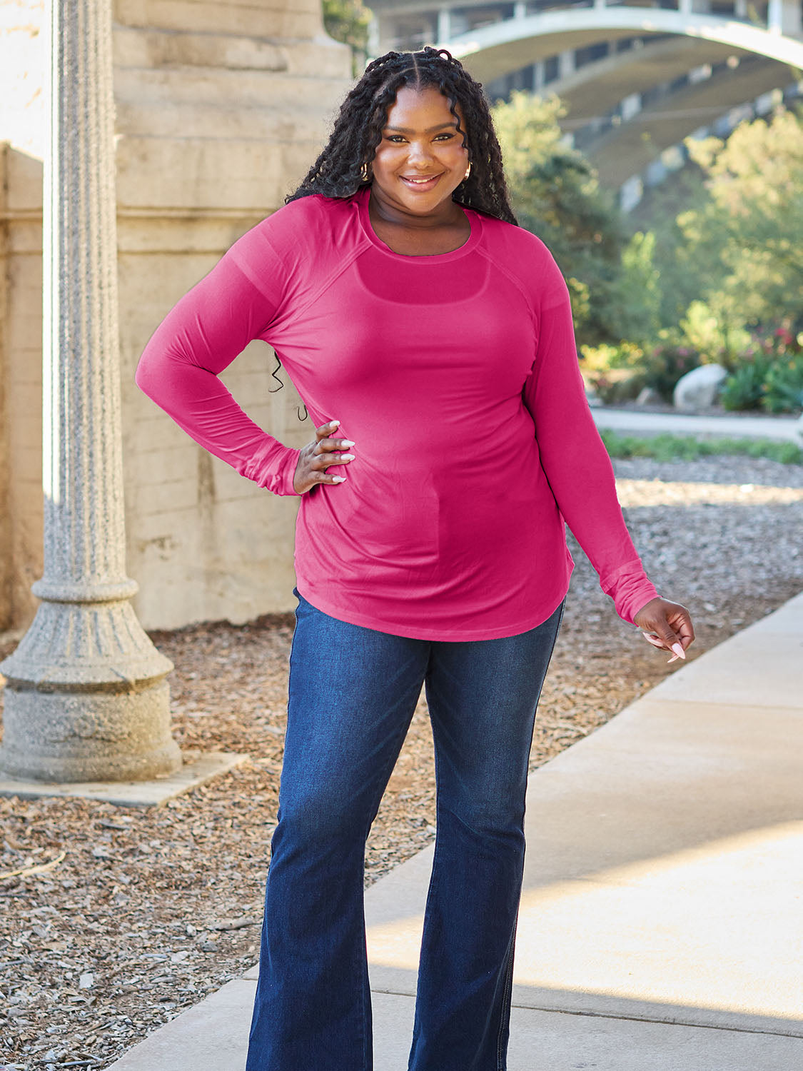 A woman wearing sunglasses and a slightly stretchy Basic Bae Full Size Round Neck Long Sleeve T-Shirt in white stands on a sidewalk with an arched structure in the background, smiling slightly.