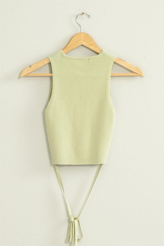 The Good Times Ribbed Sweater Tank Top, featuring a light green color, cropped hem, and back tie, hangs on a wooden hanger against a beige wall.