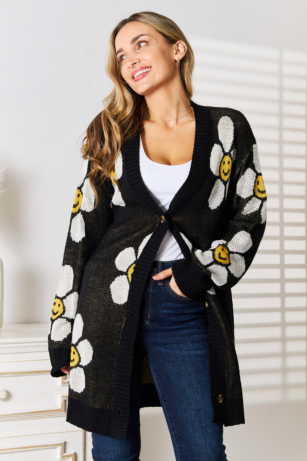 A woman stands indoors wearing the Perfee Floral Button Down Longline Cardigan, which features stylish black fabric adorned with white and yellow smiley face flowers. She pairs it with a white top and blue jeans, looking off to the side while smiling.