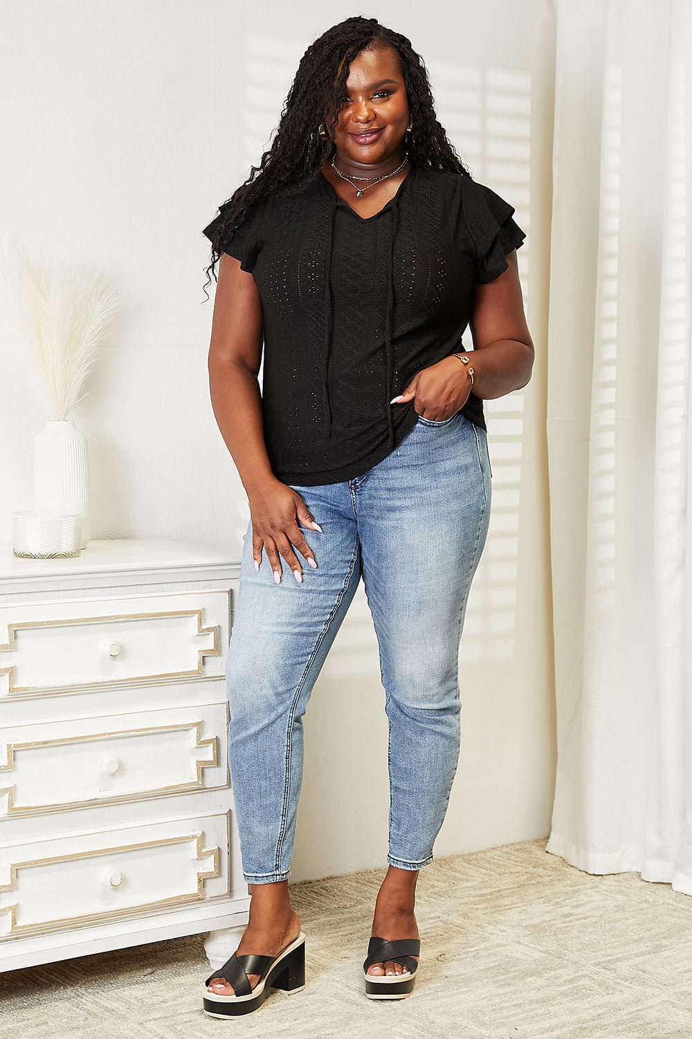 A woman with long hair wears the Mandy Eyelet Tie-Neck Flutter Sleeve Blouse and jeans, standing indoors beside a white vase with dried plants. She has her hand resting on her hip and is slightly smiling, looking off to the side.