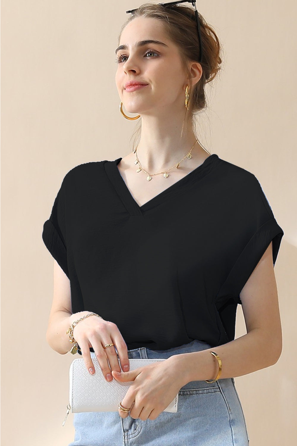 A woman wearing a stylish Ninexis V-Neck Trim Rolled Short Sleeve Shirt in light blue and high-waisted blue jeans, accessorized with hoop earrings, a necklace, and sunglasses on her head, poses in front of a beige background.