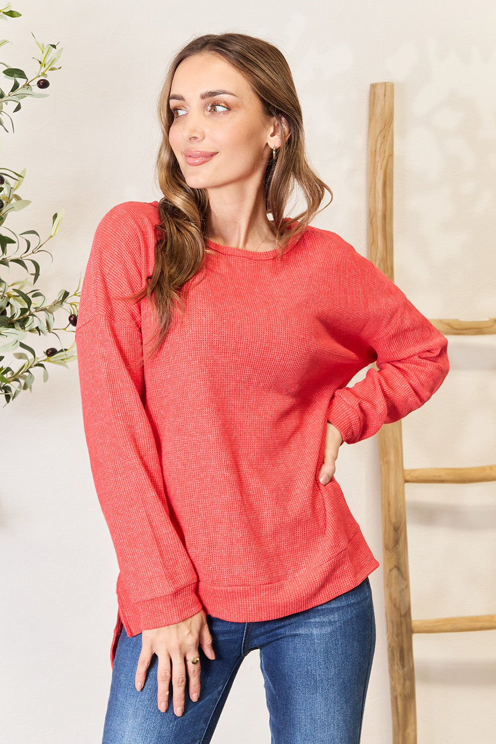 A woman wearing the Basic Bae Round Neck Drop Shoulder Slit Sweatshirt in red and blue jeans stands indoors, smiling and touching her hair, with a decorative plant and wooden ladder in the background.