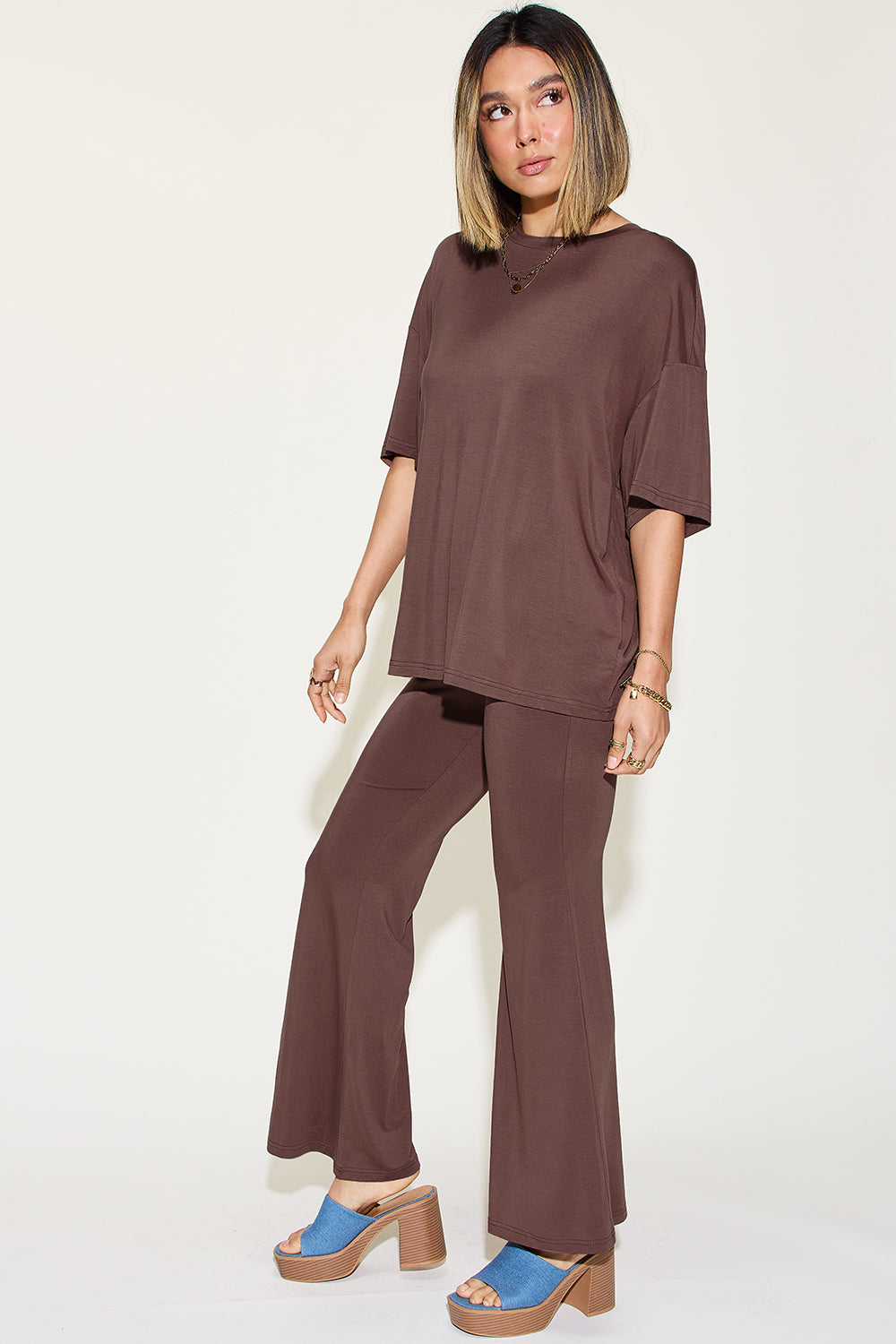 A woman with long, blonde hair wears the Basic Bae Full Size Bamboo Drop Shoulder T-Shirt and Flare Pants Set in black, paired with brown heeled sandals and layered necklaces. She stands against a plain, light background.