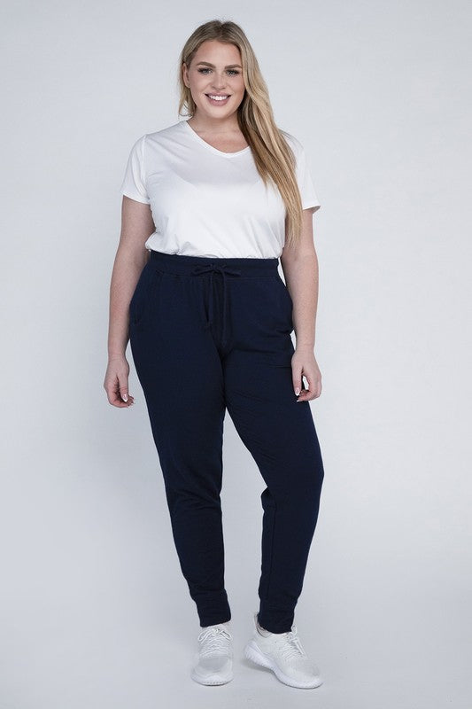 A person stands against a white background, wearing a white t-shirt, grey Plus-Size Jogger Pants with an adjustable waistband, and white sneakers. They have long blonde hair and are posing with one hand on their hip.