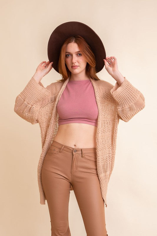 A person with long hair, wearing an oversized Knit Netted Cardigan, a crop top, and denim shorts, stands in a light-colored room.