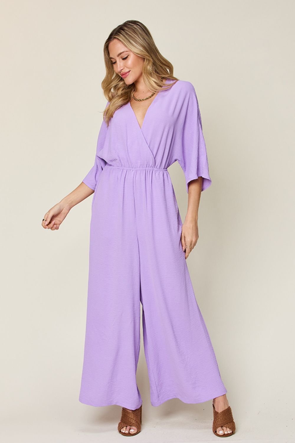 Person wearing the Double Take Full Size Surplice Wide Leg Jumpsuit with Pockets in royal blue, featuring wide legs, a deep V-neckline, and ruched detailing, posing with hands on hips against a plain light-colored background.