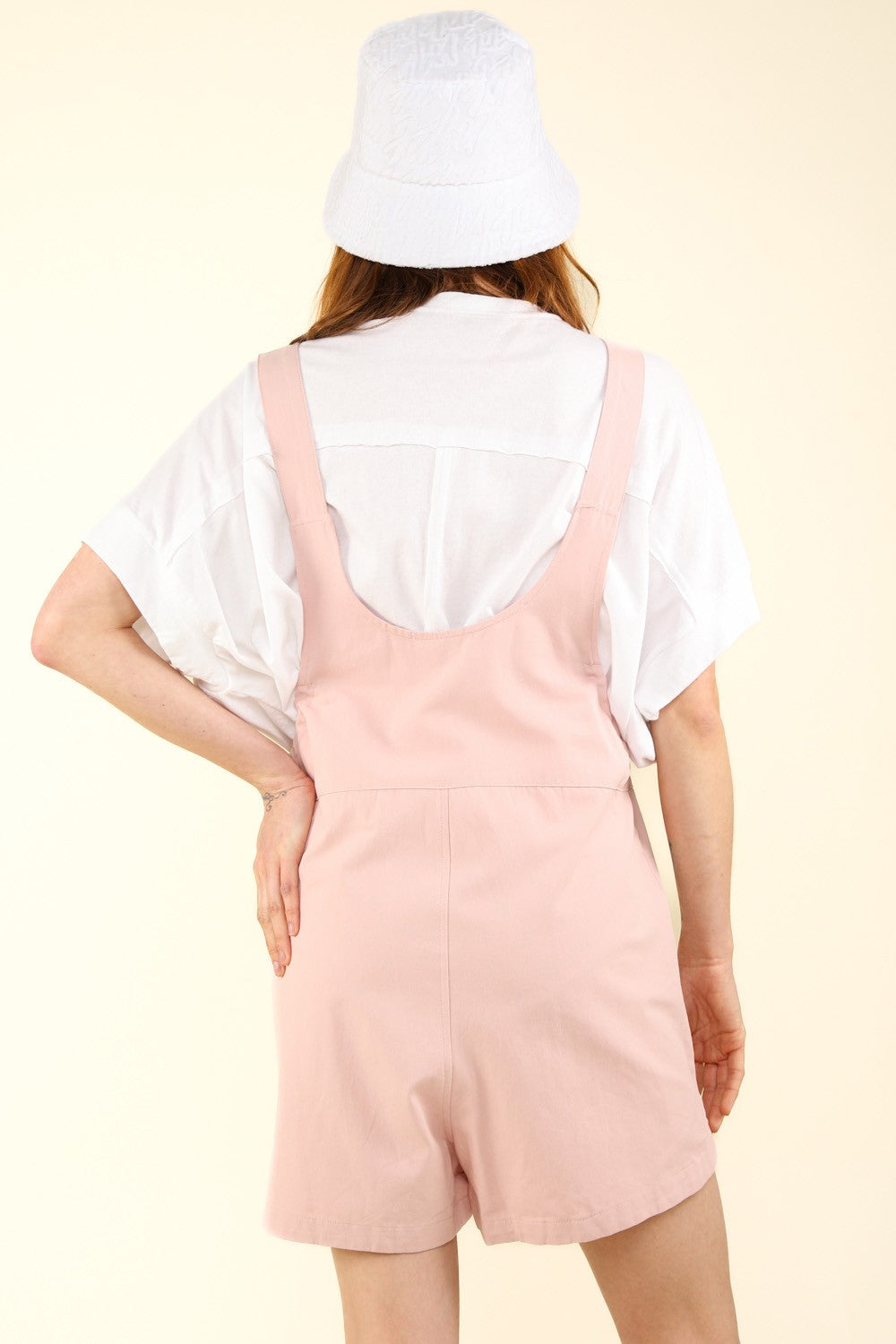 Person wearing a white hat, a white T-shirt, and VERY J Adjustable Waist Suspender Overalls with Pockets in light pink, poses with hands in the side pockets against a plain background.