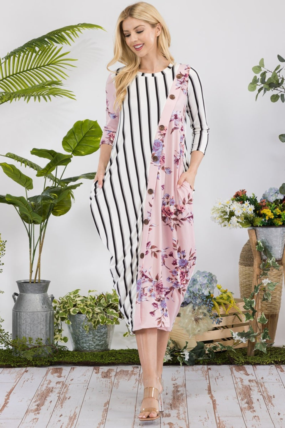 A woman stands amidst potted plants and flowers on a wooden floor, wearing the elegant and chic Celeste Full Size Floral Striped Contrast Midi-Dress with Pockets.