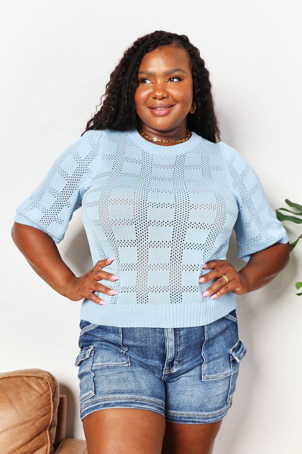 A person is standing and smiling, wearing the Mandy Ribbed Trim Round Neck Knit Top in light blue with half sleeves, paired with blue jeans. A potted plant is partly visible next to them against the plain background.