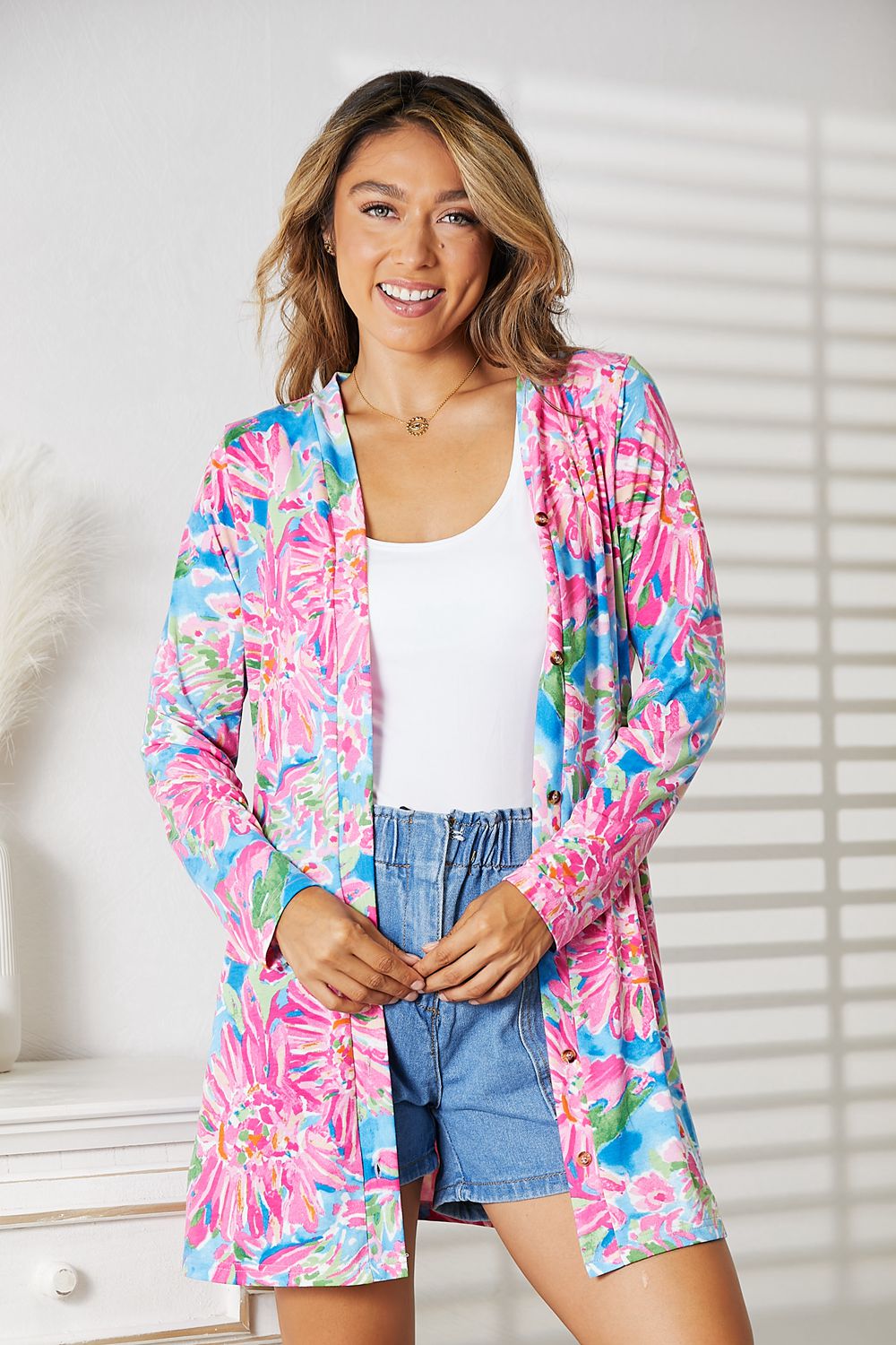 Wearing the Double Take Floral Open Front Long Sleeve Cardigan in shades of pink and blue over a white top and denim shorts, a person exudes feminine charm while standing in a well-lit room, smiling.