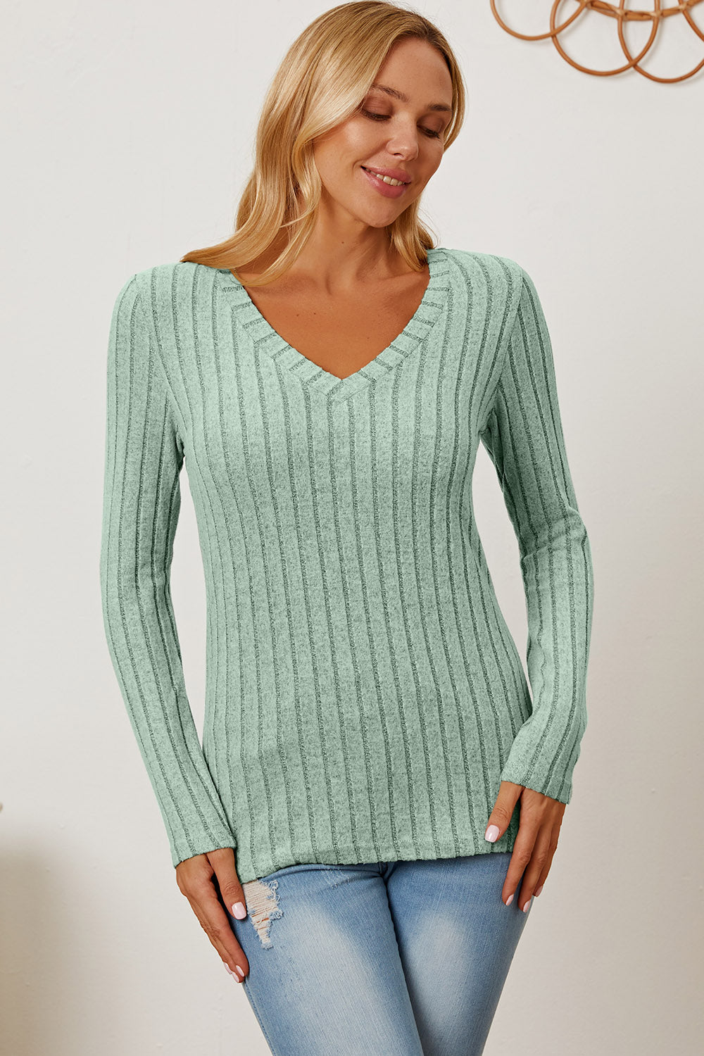 A blonde-haired woman smiles while wearing the Basic Bae Full Size Ribbed V-Neck Long Sleeve T-Shirt, which is light green and slightly stretchy, paired with light blue jeans. She holds one hand near her head and the other near her waist.