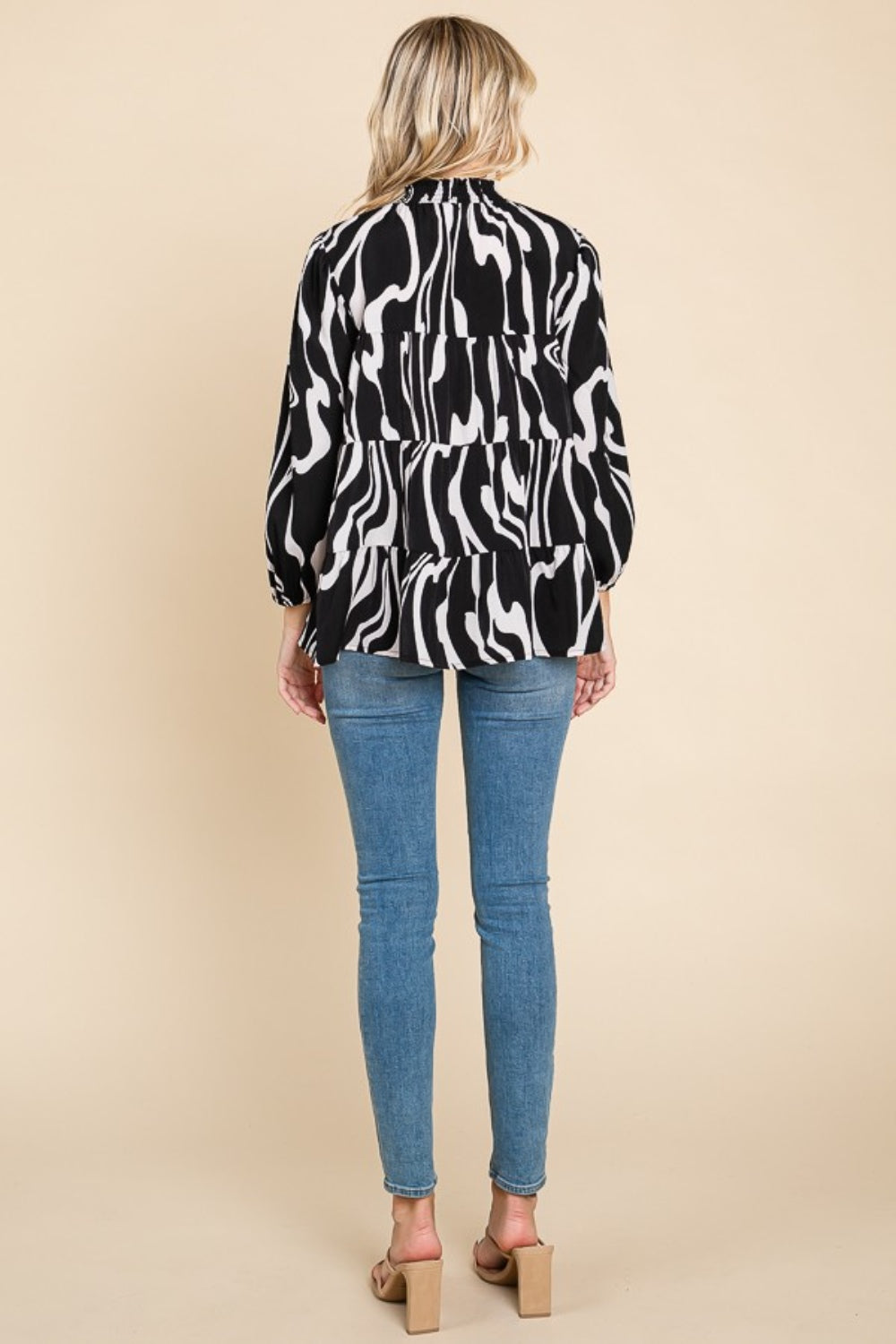 A person wearing a Culture Code Printed Smock Neck Tiered Blouse, featuring a black and white patterned design and long sleeves, paired with blue jeans, stands against a beige background.