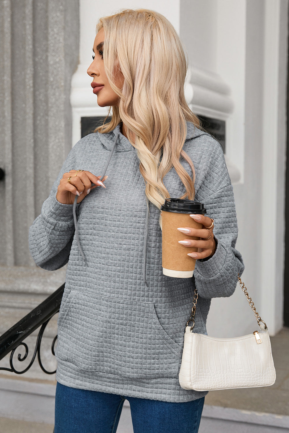 A person with long blonde hair is seen from behind, wearing a Gray Quilted Kangaroo Pocket Drawstring Hoodie and blue jeans. The relaxed fit of the textured fabric gives a casual vibe as they stand outdoors near a staircase with a metal railing.