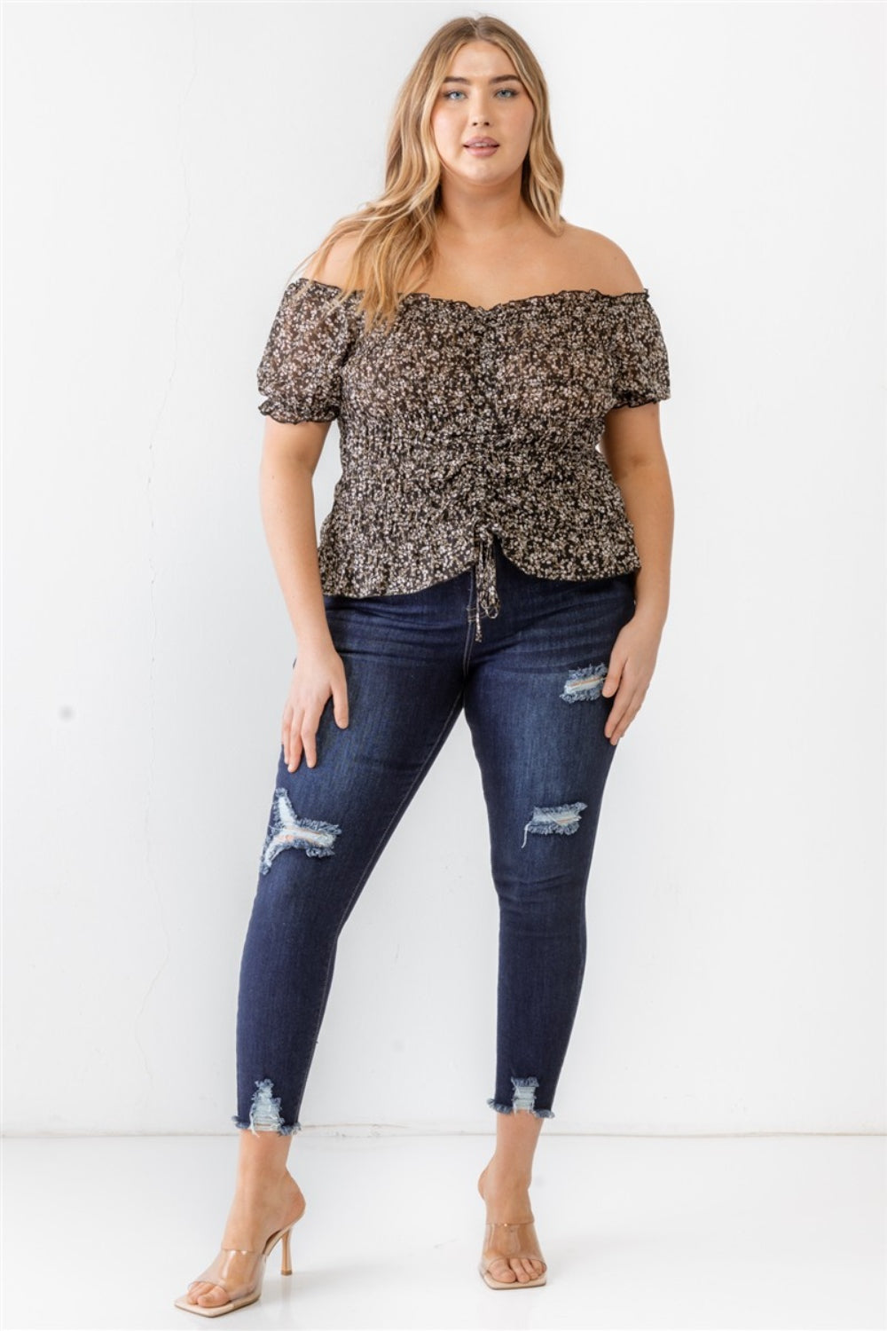 Wearing the Zenobia Plus Size Frill Ruched Off-Shoulder Short Sleeve Blouse and ripped jeans, a person with long hair poses against a plain white background.