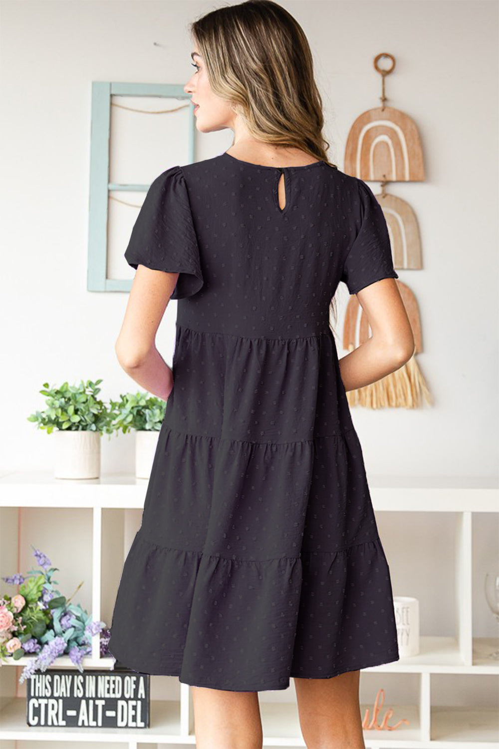 A woman elegantly poses indoors wearing the Heimish Swiss Dot Short Sleeve Tiered Dress in chic black, showcasing its stylish tiered design. The lush plants and tidy shelves create a perfect backdrop for her ensemble.