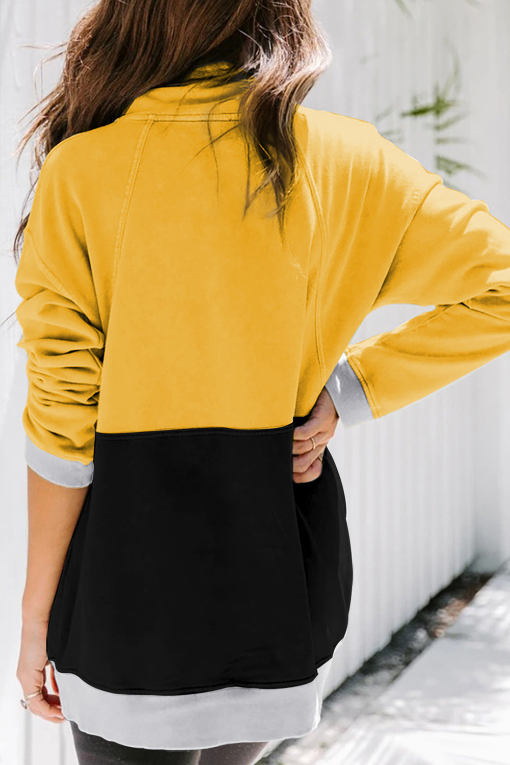 A person with long, light hair in a ponytail, wearing a Black Zipped Colorblock Sweatshirt with Pockets, blue jeans, and glasses, is holding a white cup and a brown handbag, shown from behind against a gray wall.