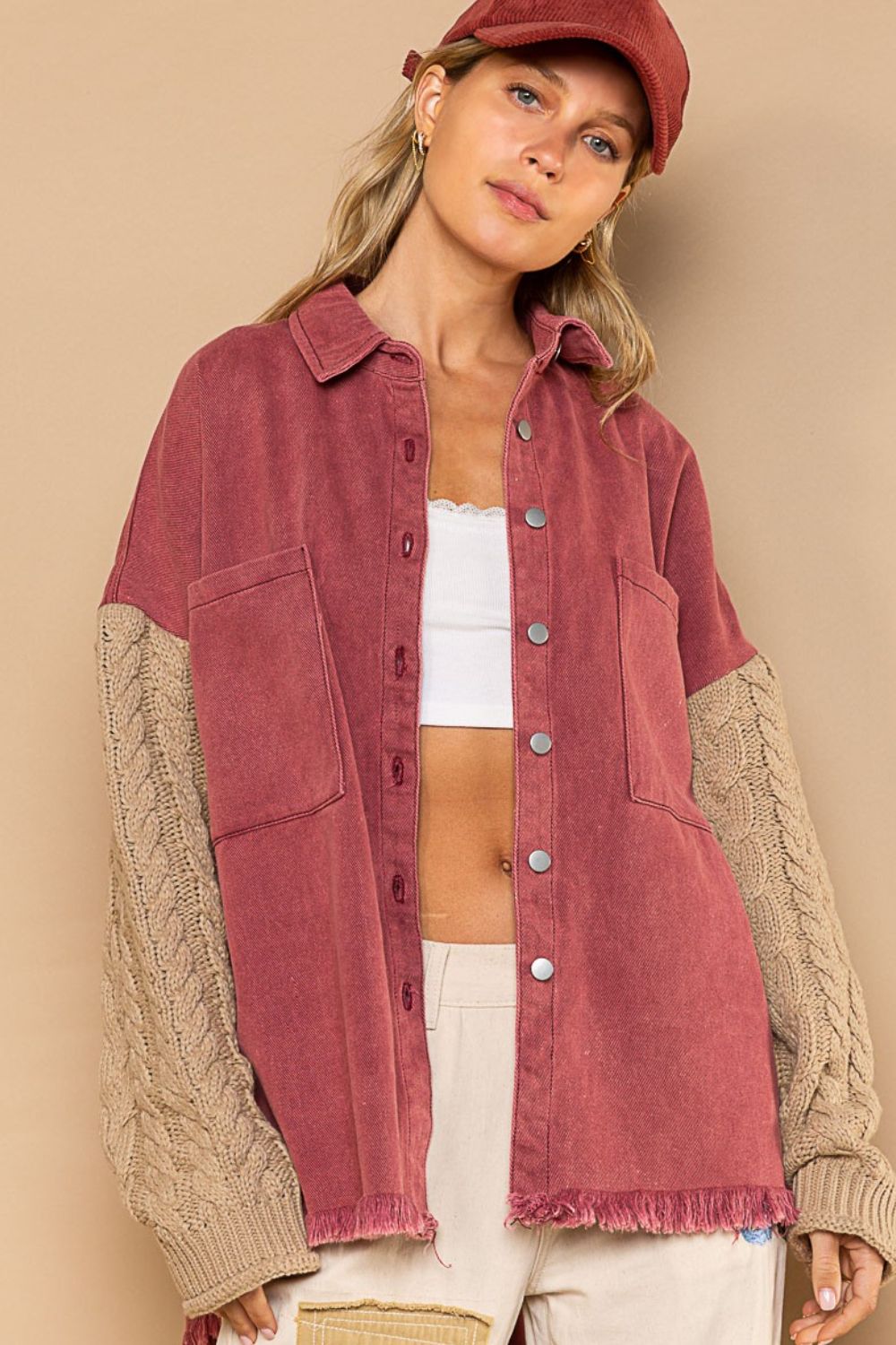 A woman wearing a POL Contrast Sweater Sleeve Button Down Shacket with beige cable-knit sleeves, a white crop top, beige pants, and a red cap poses against a neutral background.