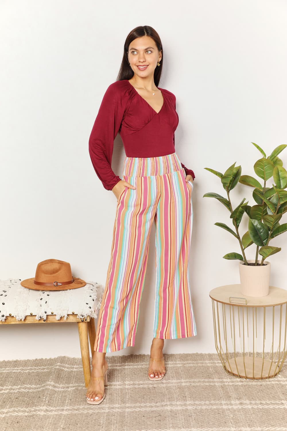 A person wearing Double Take Striped Smocked Waist Pants with Pockets and a burgundy long-sleeve shirt stands beside a wooden stool with a hat on it.