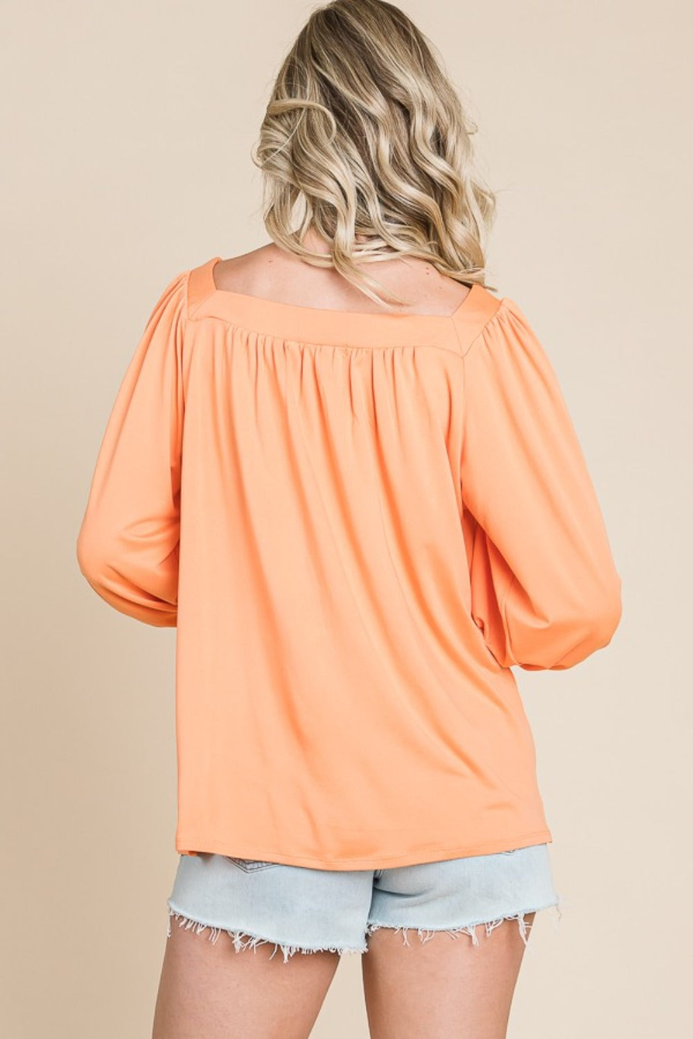 A person with long blonde hair is smiling and wearing the trendy Culture Code Square Neck Puff Sleeve Top in a light orange hue, paired with light denim shorts, showcasing a versatile fashion piece against a beige background.