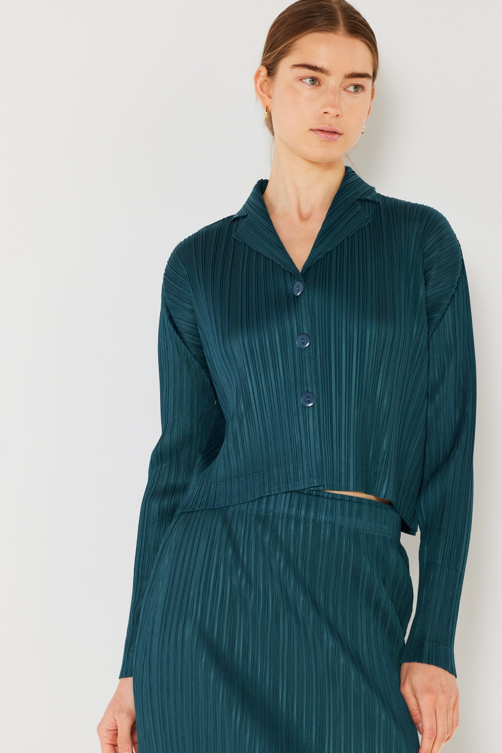 A person wearing the Marina West Swim Pleated Cropped Button Up Shirt in teal, complete with long sleeves and a matching skirt, stands against a light background, adding a modern touch to the classic pleats.