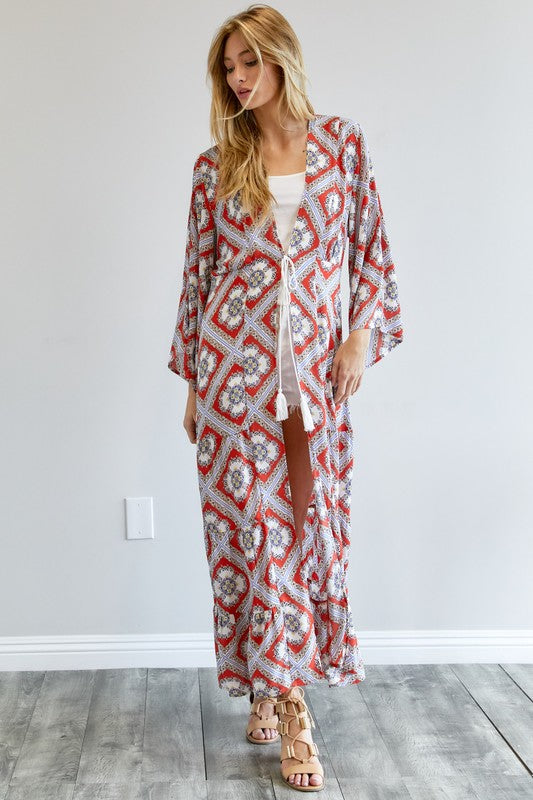 A person wearing a Printed Long Sleeve Loose Kimono with a bohemian blue and pink print over a white outfit stands in a room with a gray floor and white wall.