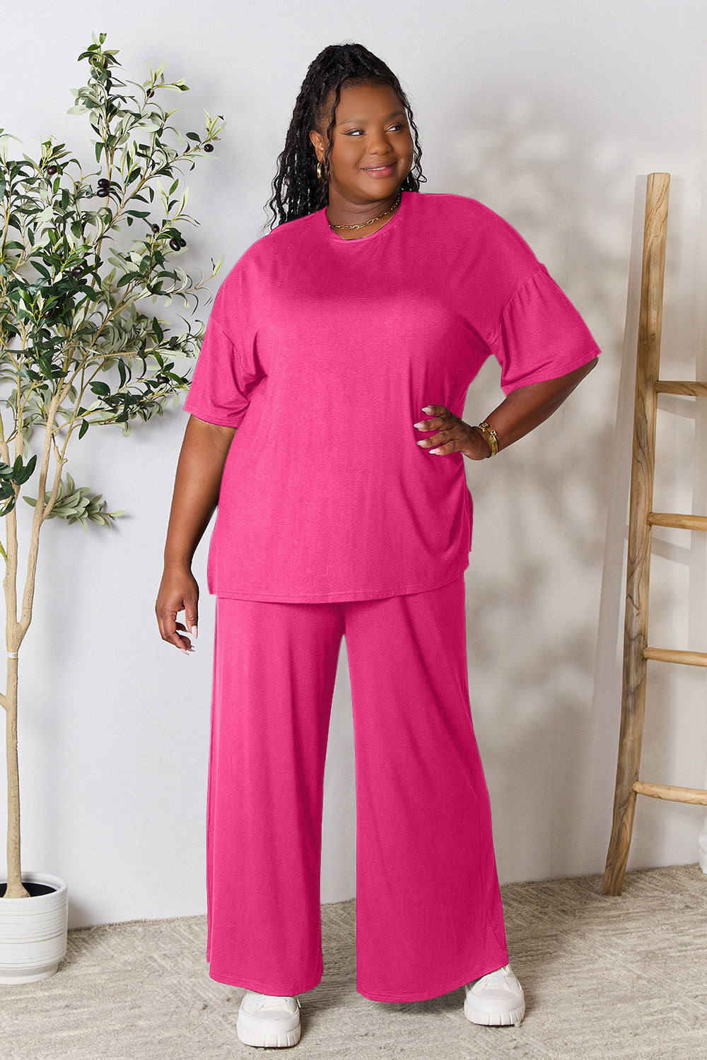 A woman stands near a potted plant and wooden ladder, wearing the Double Take Full Size Round Neck Slit Top and Pants Set in a bright pink two-piece outfit with matching wide-leg pants that are available in plus size and are easily machine washable.