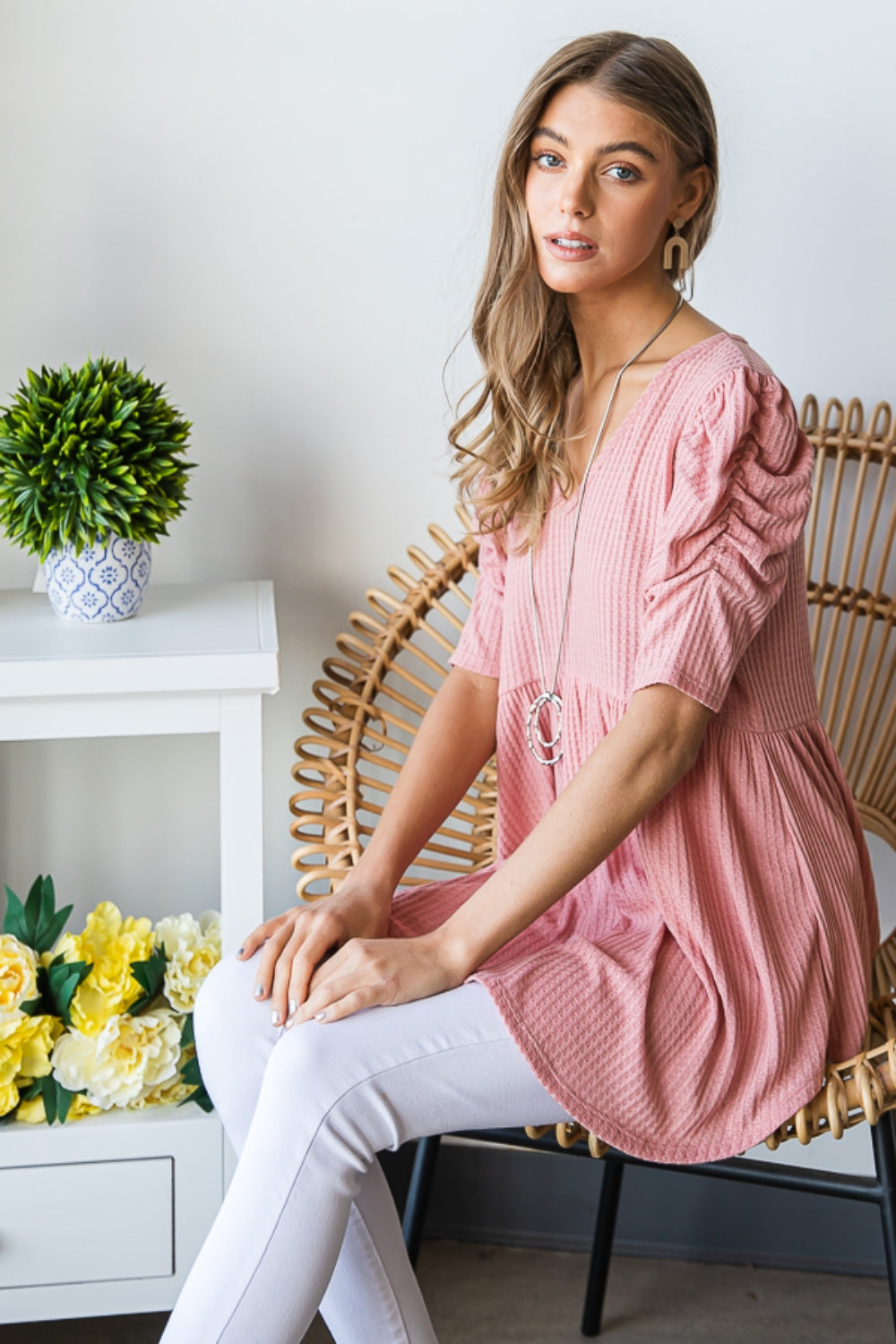 A woman in a Heimish Full Size Waffle Knit V-Neck Babydoll Top with short-sleeves and a long necklace, standing by a window with a neutral expression; the perfect cozy wardrobe addition.