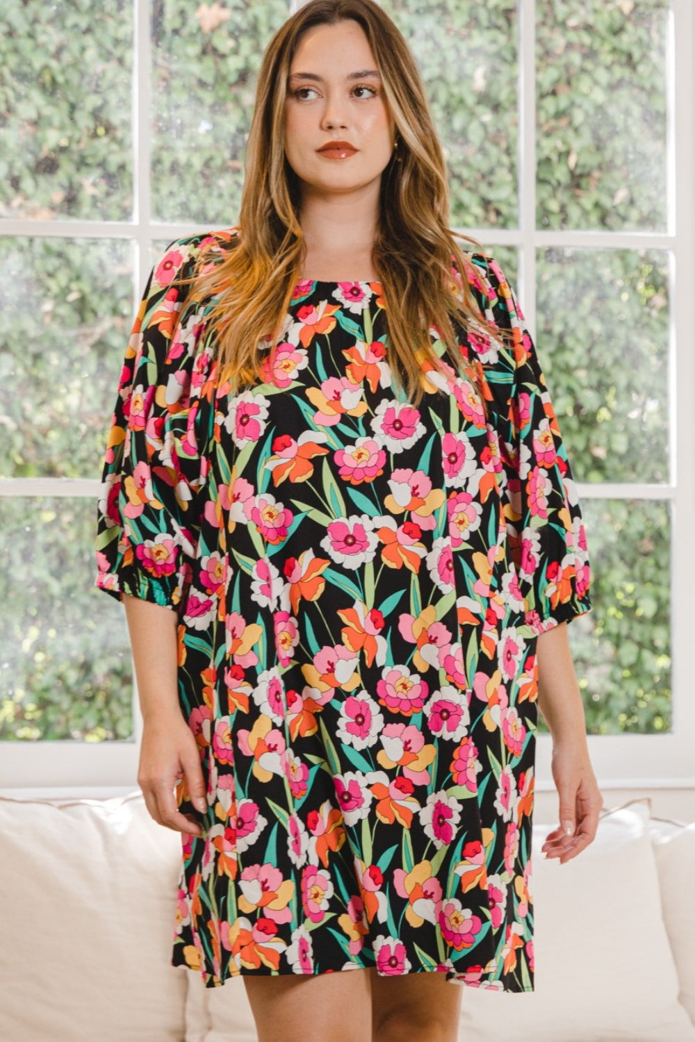 A woman wearing the stylish ODDI Full Size Floral Puff Sleeve Mini Dress stands indoors, framed by a large window with lush greenery outside.