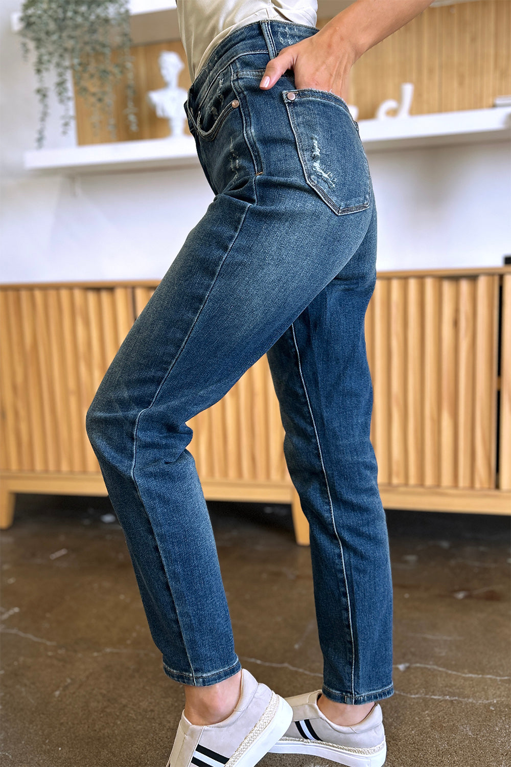 Person wearing Judy Blue Full Size Tummy Control High Waist Slim Jeans in blue and a white shirt, with their hands in their pockets. They are standing indoors on a polished concrete floor with a wooden cabinet and plant in the background.