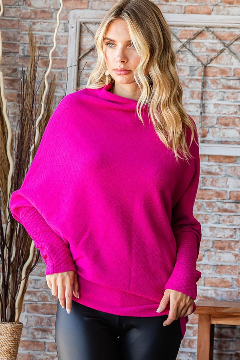 A person with long, blonde hair is standing indoors, smiling, and wearing a bright pink First Love Full Size Asymmetrical Hem Dolman Sleeve Sweater paired with black pants.