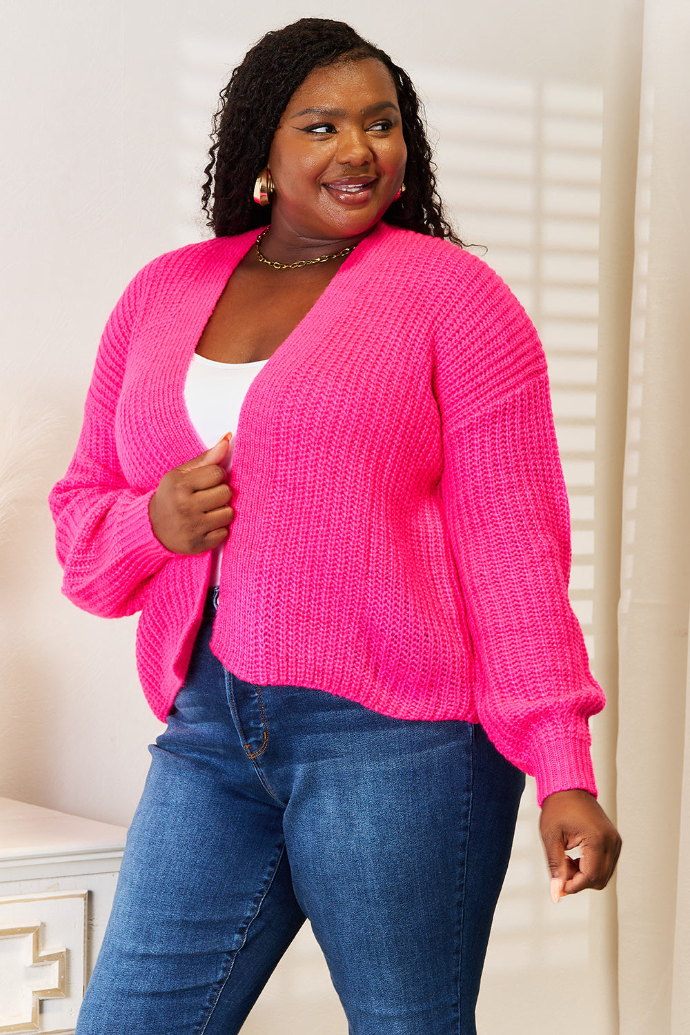 A woman wearing the Woven Right Rib-Knit Open Front Drop Shoulder Cardigan in bright pink over a white top and blue jeans stands indoors, looking to the side.