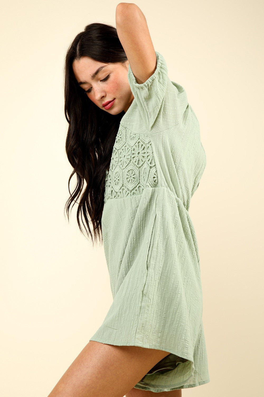 A person with long dark hair wearing a VERY J Lace Detail Puff Sleeve Romper with Pockets in light green, featuring a patterned bodice, stands against a plain beige background.