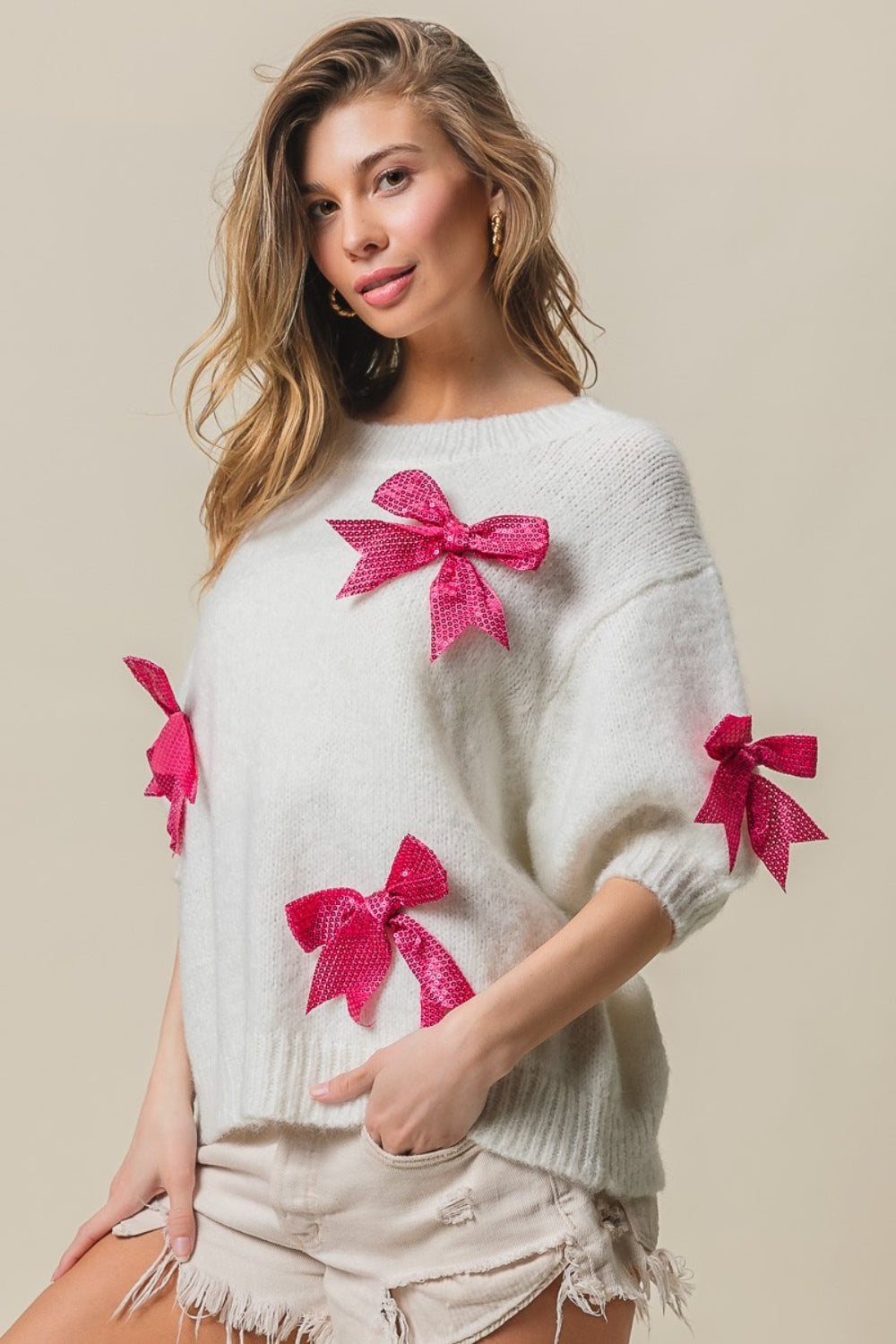 A woman wearing a striking and fashionable BiBi Sequin Bow Puff Sleeve Sweater, paired with light-colored shorts, stands against a plain background.