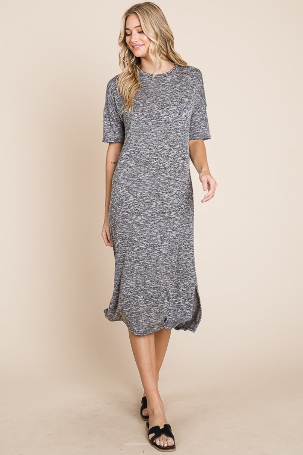 A woman wearing the BOMBOM Slit Round Neck Half Sleeve Midi Dress, a knee-length, grey midi dress featuring short sleeves and a slit detail at the hem, paired with black slip-on sandals, stands in front of a neutral background.