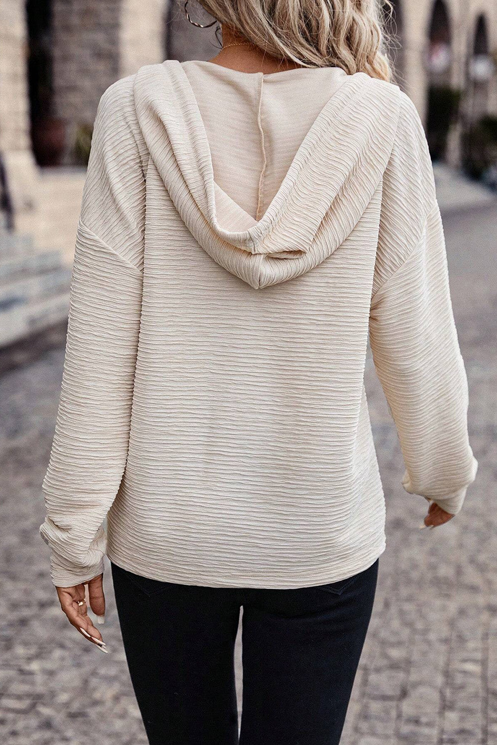 A person is wearing an Apricot Solid Color Textured Drawstring Henley Hoodie and black pants. They are holding a beverage cup and have rings on their fingers. Their face is not visible, making it perfect for casual occasions.