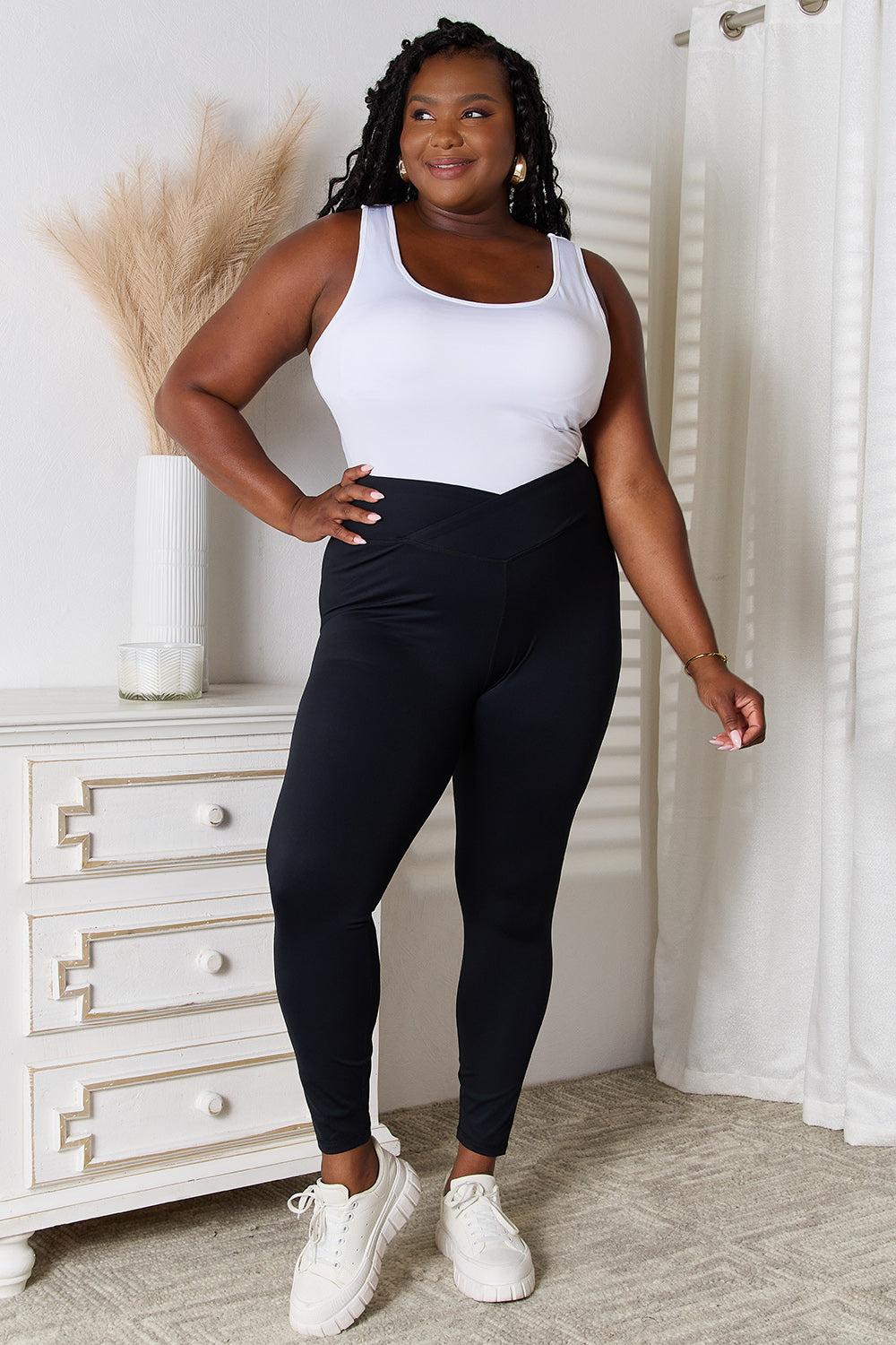 A person is standing in a room wearing Basic Bae V-Waistband Sports Leggings that offer a flattering fit, paired with white slip-on shoes. To the left, there is a white dresser with drawers. This outfit highlights an active lifestyle, seamlessly blending comfort and style.