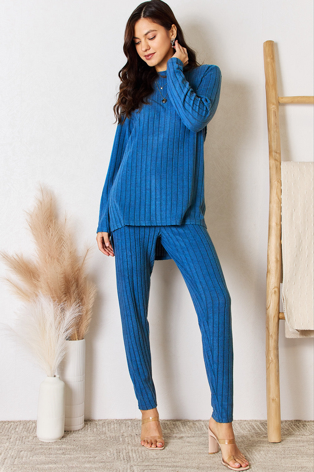 A woman in a Basic Bae Full Size Ribbed Round Neck High-Low Slit Top and Pants Set poses indoors near a wooden ladder. She is wearing the matching maroon slightly stretchy top and pants, paired with beige heels. Pampas grass in a white vase is visible in the foreground. For care, machine wash cold.