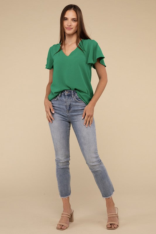 A person stands against a beige background, wearing a green Woven Airflow Flutter Sleeve Top with chic breathable fabric and high-waisted blue jeans. Hands are in their pockets, and they have long brown hair.