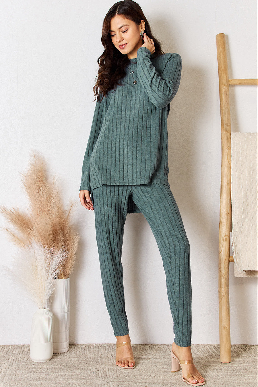 A woman in a Basic Bae Full Size Ribbed Round Neck High-Low Slit Top and Pants Set poses indoors near a wooden ladder. She is wearing the matching maroon slightly stretchy top and pants, paired with beige heels. Pampas grass in a white vase is visible in the foreground. For care, machine wash cold.