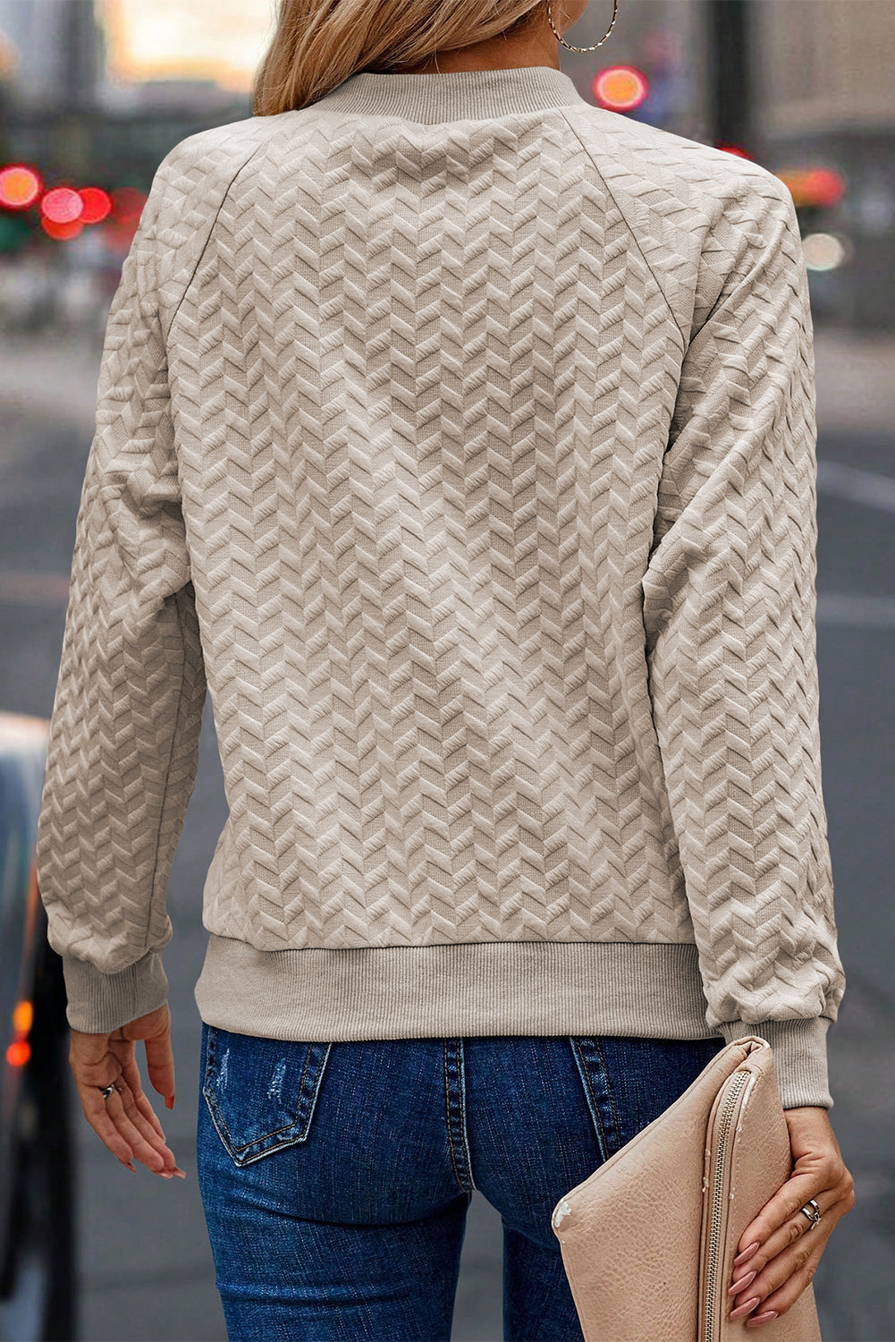 A woman in a Beige Solid Textured Raglan Sleeve Pullover Sweatshirt and blue jeans is looking to the side. She sports a brown hat and earrings, exuding a casual and comfy vibe. Standing next to dried pampas grass against a light background, she seamlessly blends with her textured sweatshirt.