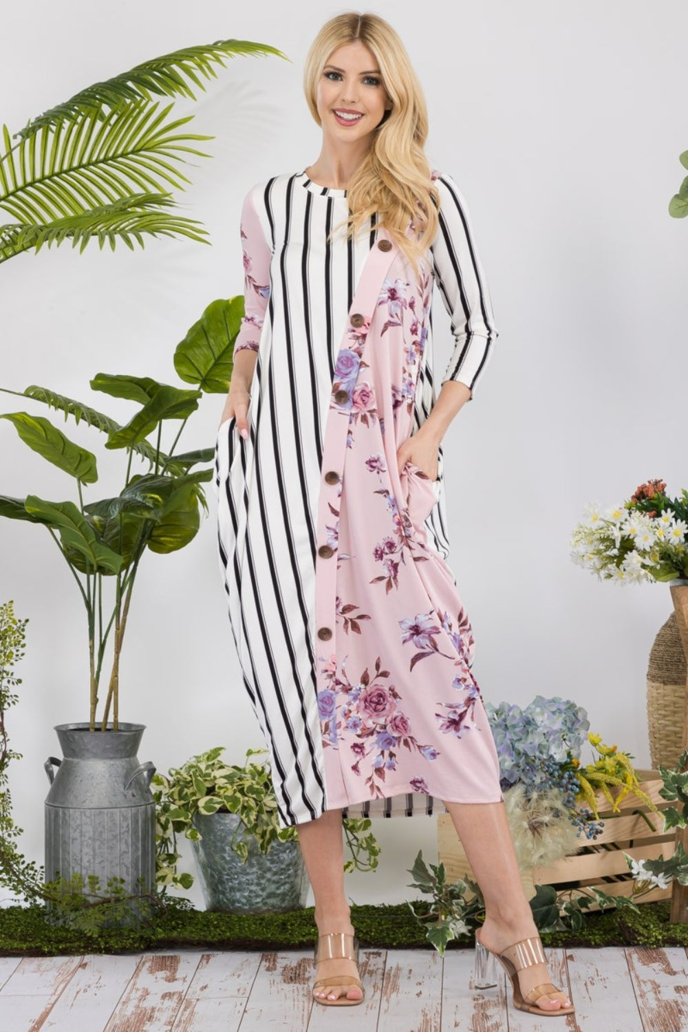 A woman stands amidst potted plants and flowers on a wooden floor, wearing the elegant and chic Celeste Full Size Floral Striped Contrast Midi-Dress with Pockets.