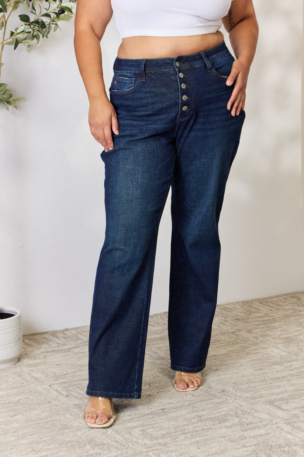 A person stands with hands in pockets, wearing the Judy Blue Full Size Button-Fly Straight Jeans, known for their high-waisted dark blue design crafted from premium lycra fabric. The jeans showcase button details and a straight leg cut, complemented by a white top and open-toed heels.